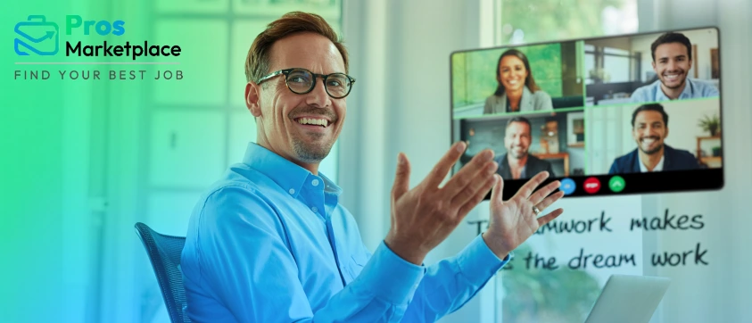 A smiling man in glasses gestures enthusiastically during a video call with four colleagues on a screen, with “Teamwork makes the dream work” written on the glass wall behind him.